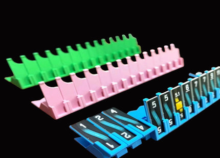Bomb Busters Player Wire Stands/ Three plastic organizer trays in green, pink, and blue, each holding numbered black cards with white text, shown on a black background.