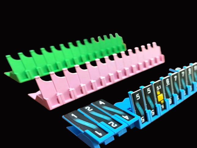 Bomb Busters Player Wire Stands/ Three plastic organizer trays in green, pink, and blue, each holding numbered black cards with white text, shown on a black background.
